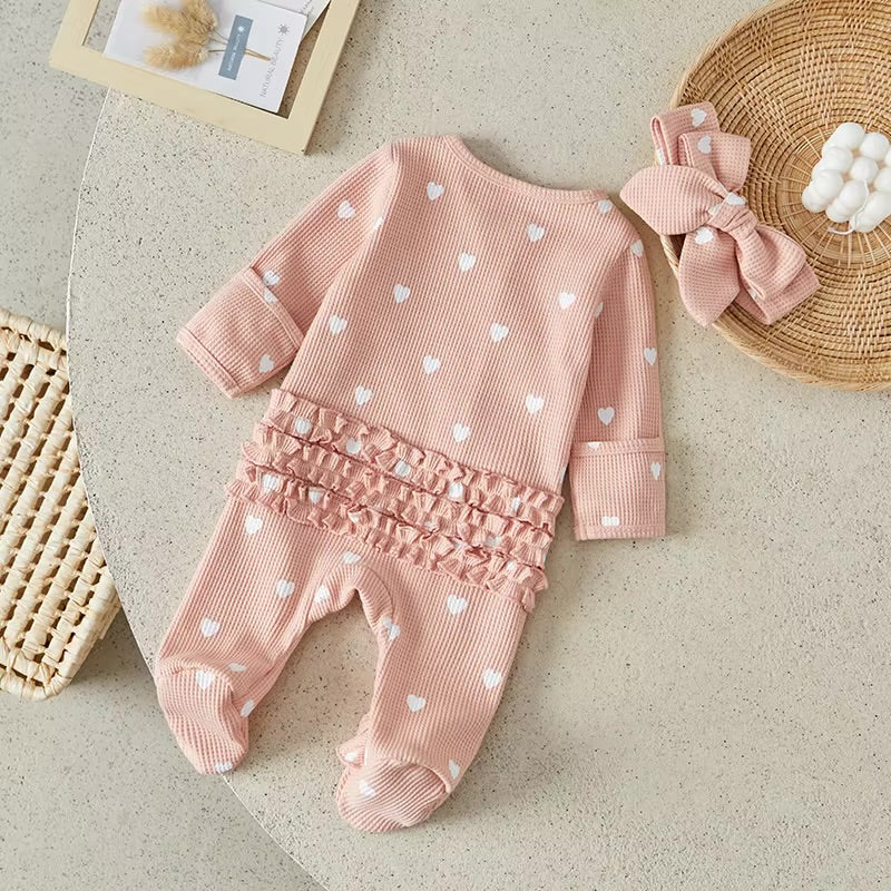 Pink baby outfit with heart patterns and matching headband on a neutral surface.