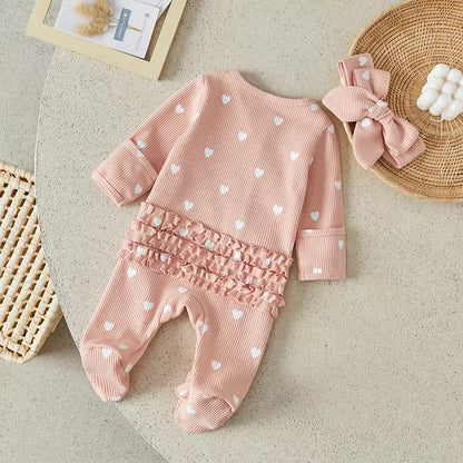 Pink baby outfit with heart patterns and matching headband on a neutral surface.