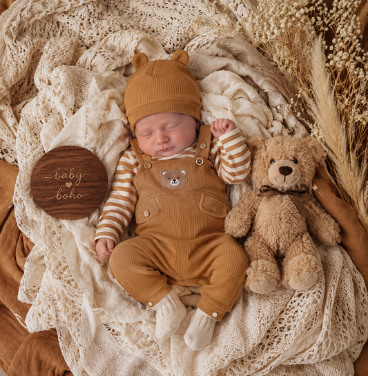 A newborn baby wearing a mustard yellow corduroy overalls outfit with a matching beanie and striped long-sleeve shirt, lying on a cream lace blanket next to a teddy bear and a round wooden sign that reads 'baby boho'.