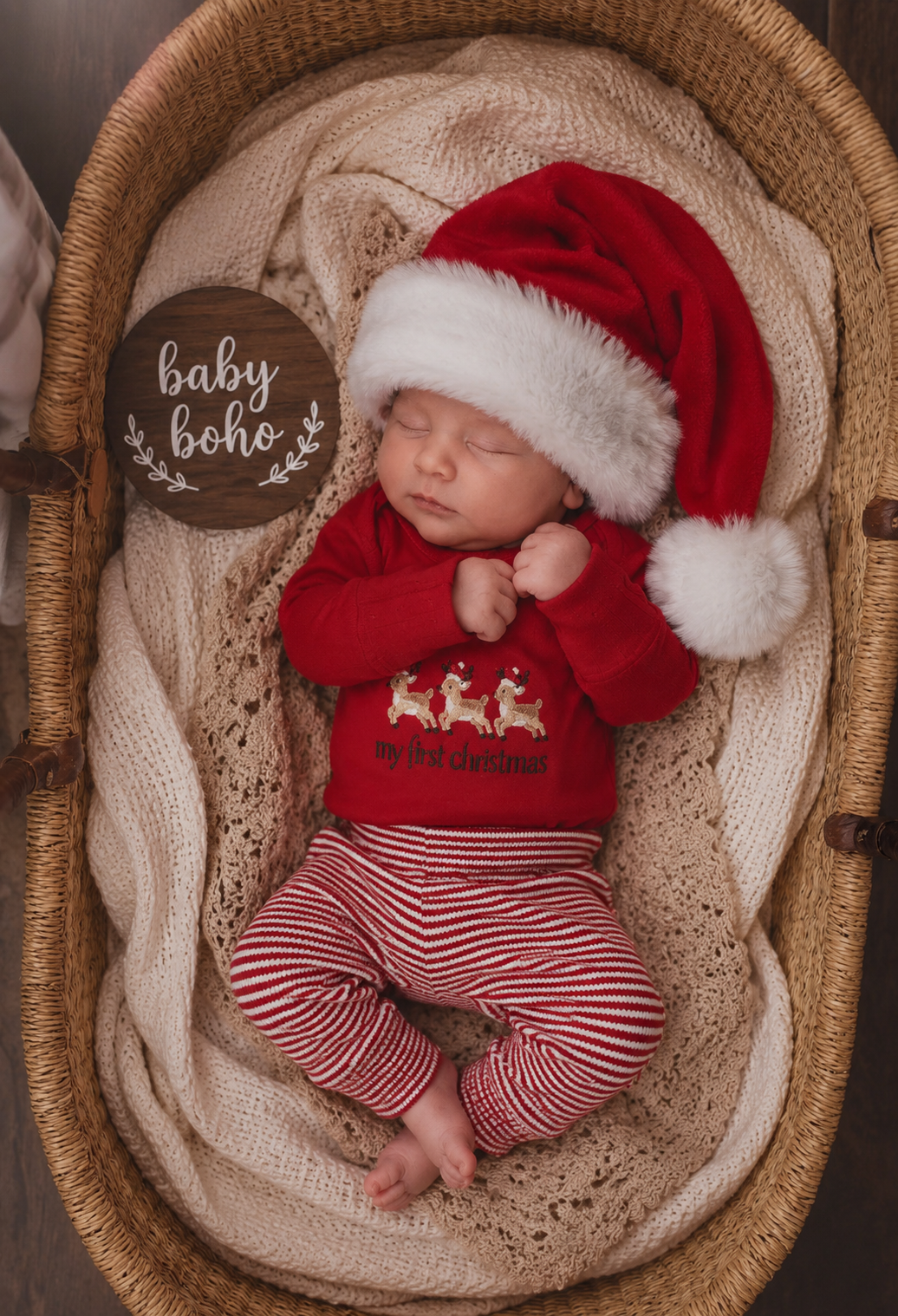 A sleeping newborn baby wearing a red Santa hat with white faux fur trim and a red long-sleeve shirt with white reindeer graphics, paired with red and white striped pants, posed inside a woven bassinet with a cream knit blanket. A round wooden sign reading 'baby boho' is placed to the left.