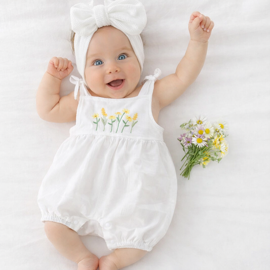 Baby wearing a white romper with floral embroidery and a matching headband, lying on a white surface with flowers beside.