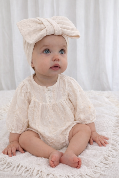 Baby wearing a cream-colored outfit and headband sitting on a white surface.