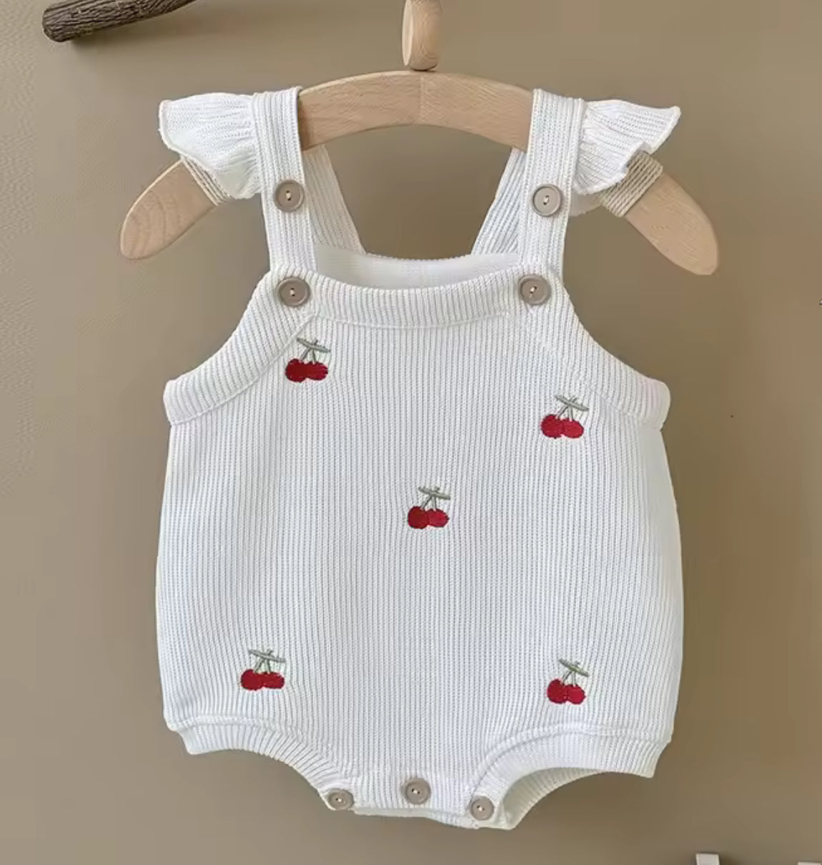 White baby romper with cherry designs on a wooden hanger against a beige background