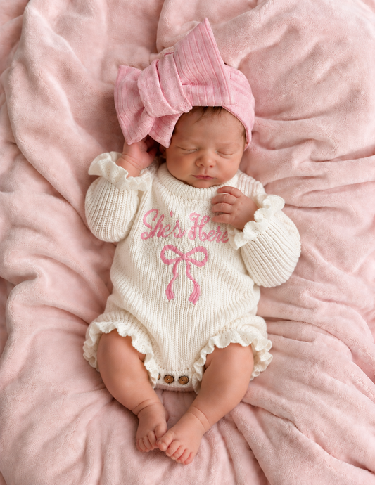Baby boho clothing cordon knit newborn romper -Newborn baby in a white outfit with pink text and bow, wearing a matching pink headband, on a pink blanket.
