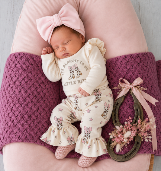 Newborn baby in a white outfit with floral patterns, pink headband, and pink knitted leg warmers.
