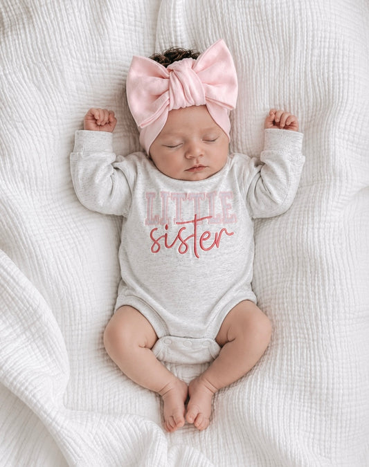 Baby boho A sleeping newborn baby wearing a light gray long-sleeve bodysuit with 'TITLE sister' printed on the front in pink, along with a matching pink bow headband, lying on a white textured muslin blanket.