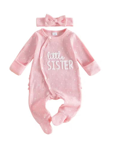 A baby girl's footed sleeper in light pink with white polka dots and 'little SISTER' text on the chest, shown with a matching pink bow headband on a white background.