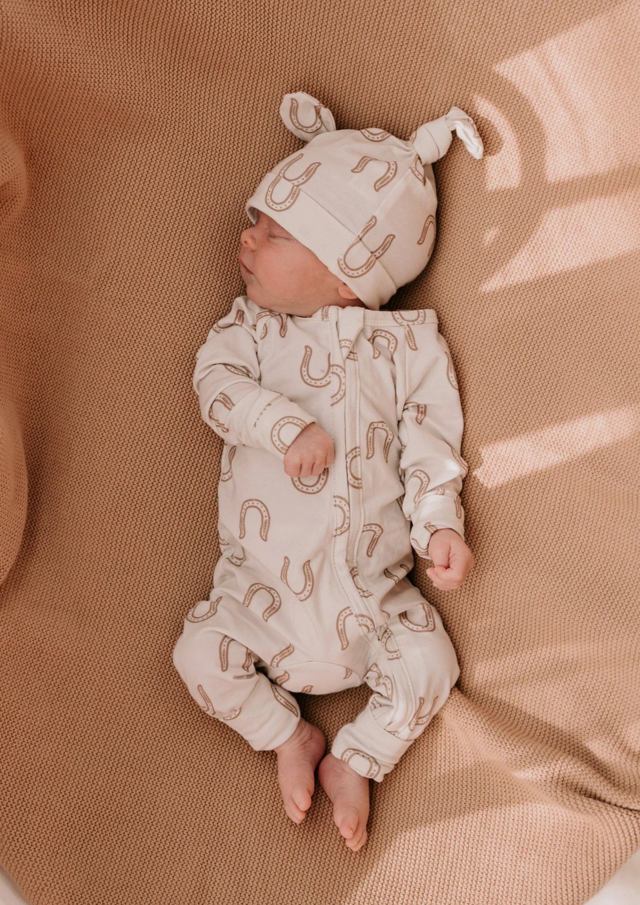 A newborn baby lying on a textured tan little and fern - get it at Baby boho - romper blanket, wearing a matching cream-colored footed sleeper and hat with a brown horseshoe pattern.