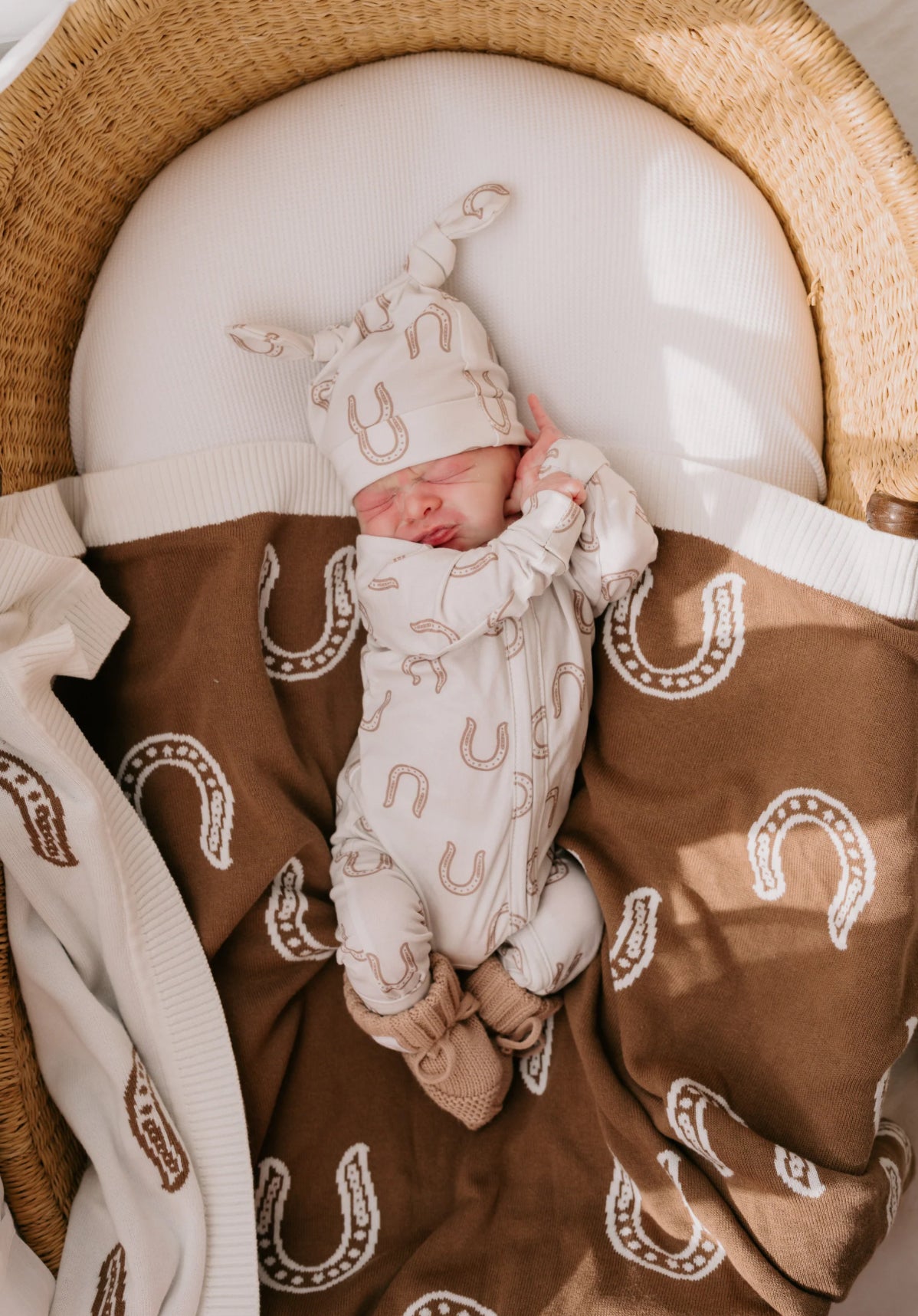 Little and fern- sold at Baby boho A newborn baby wearing a cream bodysuit and matching hat with brown horseshoe patterns, sleeping in a woven basket bassinet on a brown blanket with a matching horseshoe print.