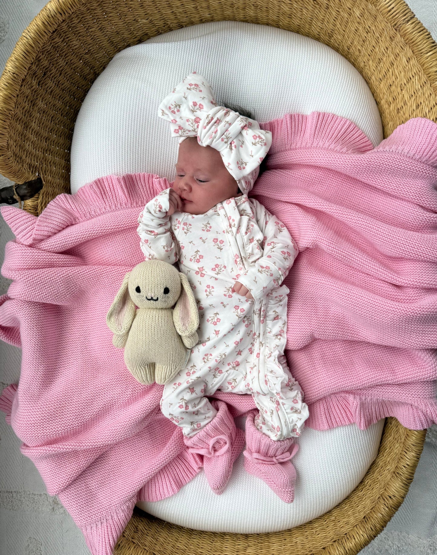 Little and fern-newborn baby wearing a white floral one-piece jumpsuit with a matching bow headband and pink booties, lying on a pink blanket inside a woven basket next to a cream stuffed bunny.Fern - at Baby Boho -